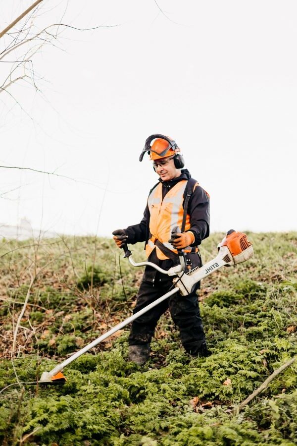 Een groenvoorziener die in de natuur staat. Hij staat in een park/bos waar geen huizen in de omgeving staan. Hij draagt werkkleding en een helm en is bezig met een machine in het gras/mos.