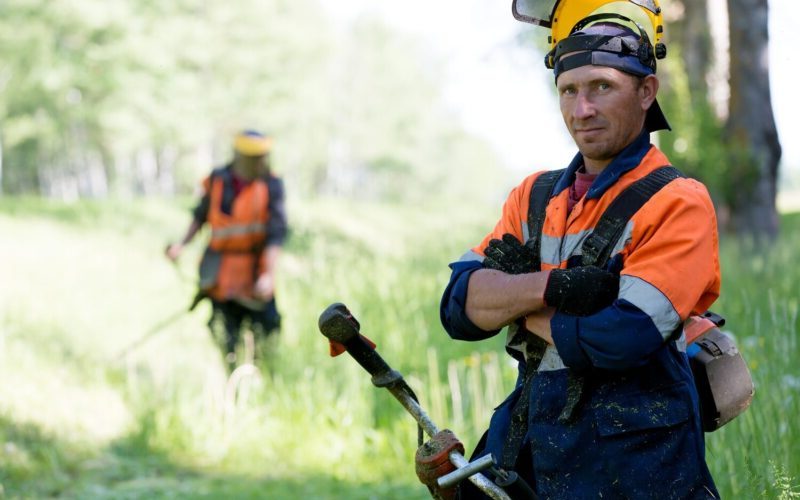 twee mannen zijn werkzaam in de groenvoorziening en zijn bezig met het maaien van gras met een bosmaaier.