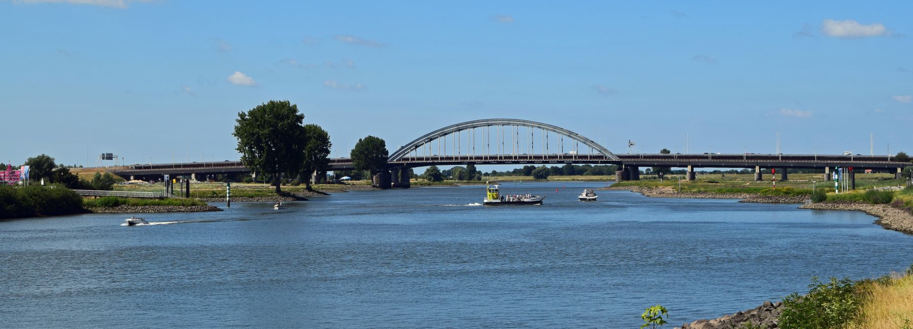 Snelwegbrug vanuit de rustige wijk met de rivier "de Lek" op de voorgrond waarop een boot en een veerboot varen.