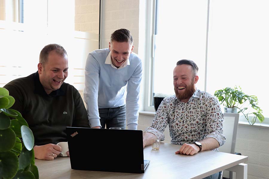 Drie mannen zijn op kantoor onder een overleg aan het lachen ten gevolge van een afbeelding die op de laptop voor hun wordt laten zien.