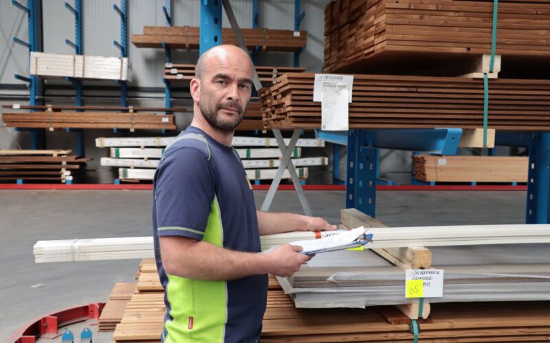 Een kale man met gezichtsbeharing werkt in een houthandel, hij was bezig met het lezen van formulieren en kijkt in de camera.